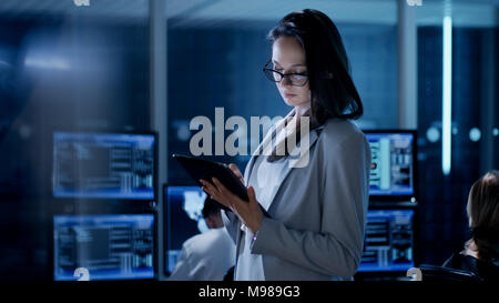 Young Female Engineer Uses Tablet in System Control Center. In the Background Her Coworkers are at Their Workspaces with many Displays Stock Photo