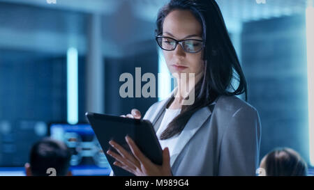 Close-up Shot of Young Female Government Employee Wearing Glasses Uses Tablet in System Control Center. In the Background Her Coworkers Stock Photo