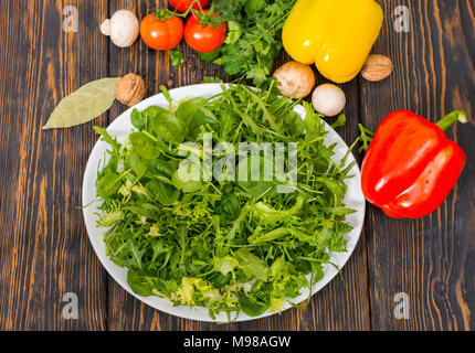 High angle view of tomatoes, red and yellow pepper, dill and parsley around white plate with mix of lettuce leaves on wooden desk Stock Photo