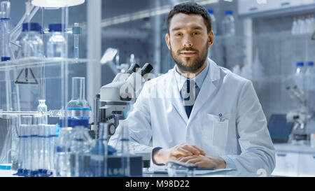Young Male Research Looks into Camera and Smiles. He's Sitting in a High-End Modern Laboratory with Beakers, Glassware, Microscope and Working Monitor Stock Photo