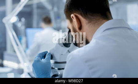 Over the Shoulder View of a Research Scientist Looking into Microscope. He's Conducts Experiments with His Colleagues in Modern Laboratory. Stock Photo