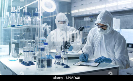 In a Secure High Level Laboratory Scientists in a Coverall Conducting a Research. Chemist Writes Down Experiment Results. Stock Photo