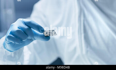 In Ultra Modern Electronic Manufacturing Factory Design Engineer in Sterile Coverall Holds Microchip with Gloves and Examines it. Stock Photo