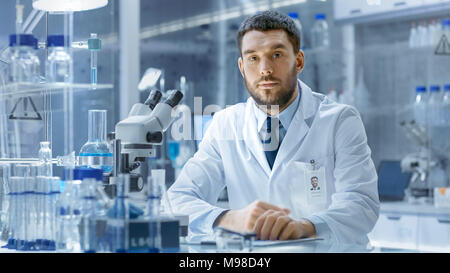 Young Male Research Scientist Talks Into Camera. He's Sitting in a High-End Modern Laboratory with Beakers, Glassware, Microscope and Working Monitors Stock Photo