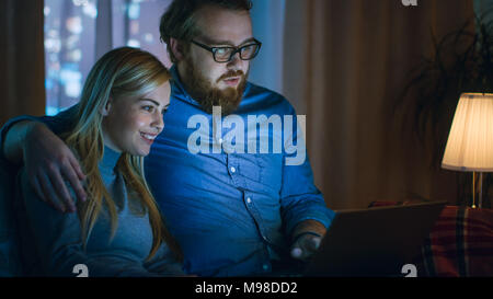 In the Evening Man and Woman are Sitting on the Sofa Watching Something on a Laptop. Living Room is Cozy. Stock Photo