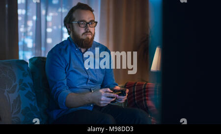 In the Evening Man is Sitting on a Sofa  and Plays in Videogames In His Living Room. Big City is Seen Behind the Windows. Stock Photo