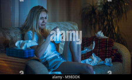 In the Evening Heartbroken Girl Sitting on a Sofa, Crying, Using Tissues, Eating Ice Cream and Watching Drama on TV. Her Room is in Mess. Stock Photo
