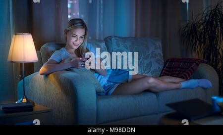 Late At Night Beautiful Woman Lying on Her Sofa in the Living Room, Using Her Smartphone and Smiling. Stock Photo