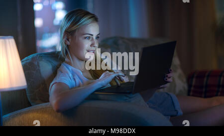 Late At Night Beautiful Woman Lying on Her Sofa in the Living Room, She Types on Her Laptop and Smiles. Her Apartment Has Big City Window View. Stock Photo