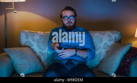 Portrait Shot of a Man Sitting on a Sofa in His Living Room, Eating Popcorn and Watching TV. Floor Lamps are Turned ON. Stock Photo