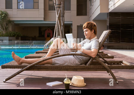 Handsome asian man relaxing by pool and reading e-book Stock Photo
