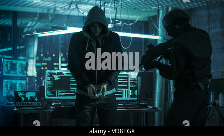 Handcuffed Hacker is Standing and Guarded by Fully Armed Special Forces Soldier. They're in Hacker's Hideout Basement with Multiple Operating Displays Stock Photo