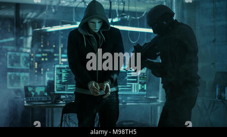 Masked Hacker Wearing Handcuffs is Guarded by Fully Armed Special Forces Soldier.  They're in Hacker's Hideout Basement with Multiple Displays Stock Photo