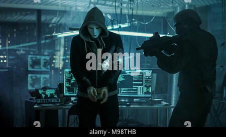 Masked Hacker Wearing Handcuffs is Guarded by Fully Armed Special Forces Soldier.  They're in Hacker's Hideout Basement with Multiple Displays Stock Photo