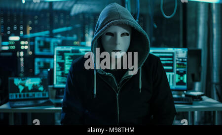 Dangerous Internationally Wanted Hacker with Covered Face Looking into the Camera. In the Background His Operating Room with Multiple Displays Stock Photo