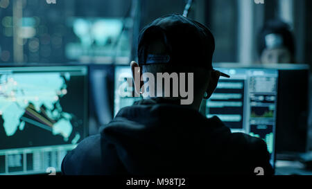 Close-up Frame from the Back of Working Hacker Organizing Advanced Virus Attack on Corporate Servers. Place is Dark and Has Multiple displays. Stock Photo