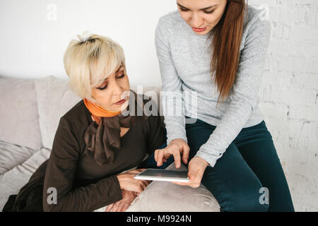 A young girl explains to an elderly woman how to use a tablet or shows some application or teaches you how to use a social network. Teaching the older generation of new technologies. Stock Photo