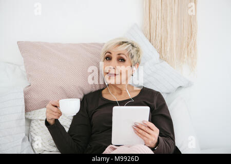 A positive woman drinks a morning coffee lying in bed and uses a tablet to watch videos, listen to music and chat with friends on social networks. Stock Photo