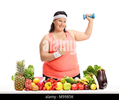 Overweight woman holding a small dumbbell and pointing behind a table with fruit and vegetables isolated on white background Stock Photo