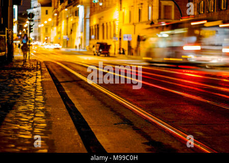 Night scene of Rome featuring a speeding bus with light trails and lights reflecting on tram lines and with motion blur and lone female figure Stock Photo