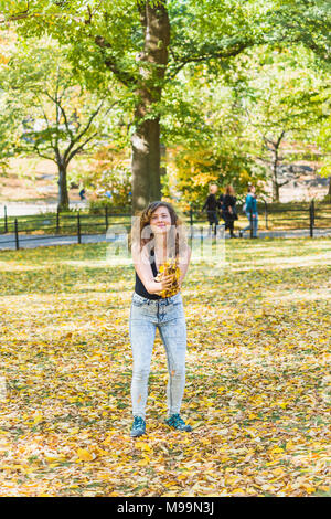 Manhattan New York City NYC Central park, young hipster millennial woman standing, throwing many fallen leaves up in air in autumn fall season with ye Stock Photo