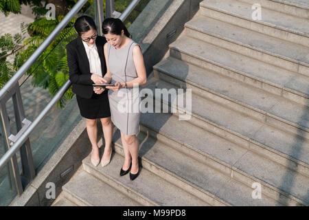 From above view.Two Asian business lady standing on the stairs using tablet discussing with each other. Stock Photo
