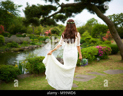 Beautiful asian woman in a white bridal dress wearing a flower crown holding a violet bouquet and showing her back in a garden with pond and pine tree Stock Photo