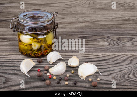 Garlic cloves pickled in oil with herbs, peppercorns and allspices. Allium sativum. Vintage jar and different varieties of spices on wood background. Stock Photo