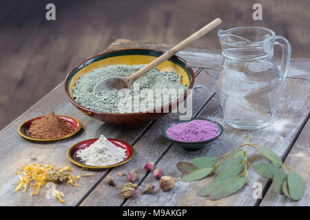 Composition of ceramic bowls of sea clay powder: red, pink, green ,purple ,pitcher of water, dryflowers, eucaliptus, concept of facial and body treatmenton wooden board, on vintage wall background  Stock Photo