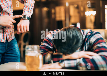 Young man in casual clothes is sleeping near the mug of beer on a table in pub, another man is waking him up. Stock Photo