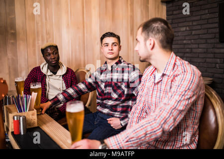 Friends met after a hard day. Three cheerful friend clink beer at the bar and drink a beer. Stock Photo