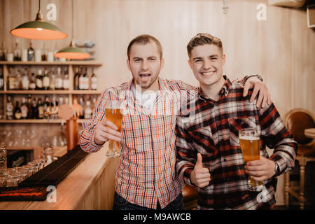 Two handsome young men in casual wear holding glasses with beer and looking away while standing at the bar counter Stock Photo