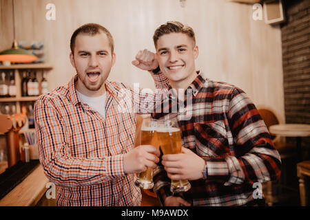 Two handsome young men in casual wear holding glasses with beer and looking away while standing at the bar counter Stock Photo