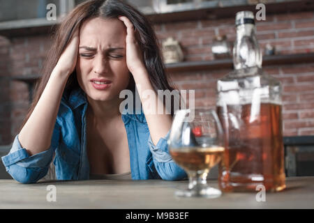 Young female alcoholic social problems sitting at table having hangover headache holding head feeling pain bottle of whiskey close-up Stock Photo