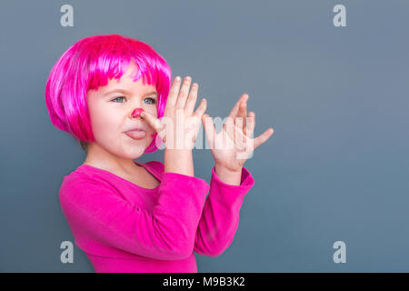 Girl in a pink suit in a wig is teased at camera. Studio shot, isolated on gray background Stock Photo