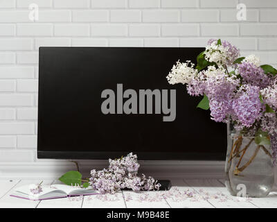 Beautiful white and violet lilac flowers bouquet in glass vase with opened note-book and black monitor screen on the white brick and wooden background Stock Photo