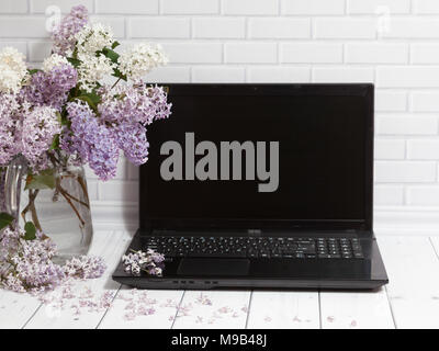 Beautiful white and violet lilac flowers bouquet in glass vase with opened black laptop on the white brick and wooden background, mock up front view Stock Photo