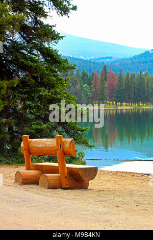 A rustic bench sits on a calm lake shore in the mountains Stock Photo