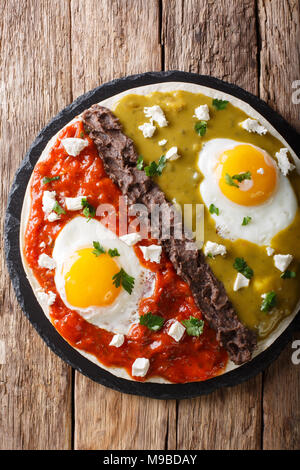 Mexican fried huevos divorciados eggs with salsa verde and roja, cheese, black beans on a tortilla close-up on a table. Vertical top view from above Stock Photo