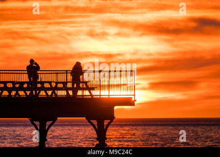 Aberystwyth Wales UK, Saturday 24 March 2018  UK Weather: People are silhouetted standing on the end of Aberystwyth pier  as the sun sets spectacularly over  Cardigan Bay ,  on the eve of British Summer Time, when the clocks go forwards and hour tonight  photo © Keith Morris  / Alamy Live News Stock Photo