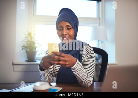 Smiling young Muslim female entrepreneur wearing a hijab sending text messages while working at table in her home office Stock Photo