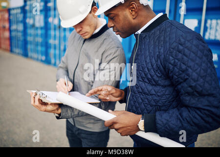 Two engineers wearing hardhats standing in a shipping container yard discussing an inventory list on a clipboard Stock Photo