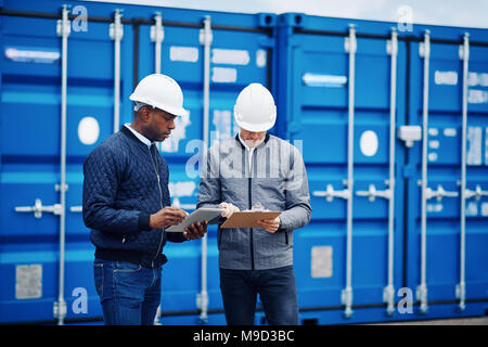 Two engineers comparing inventory lists using a tablet and a clipboard while standing by freight containers in a shipping yard Stock Photo