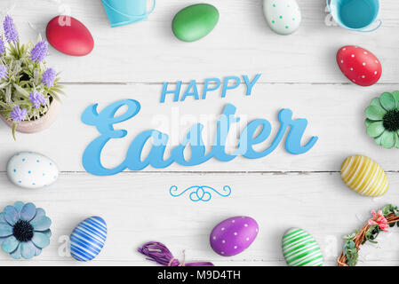 Blue happy easter text on white wooden desk surrounded with colorful eggs and floral decorations. Stock Photo