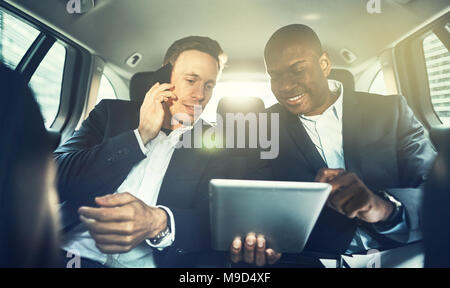 Businessman talking on his cellphone with his colleague sitting next to him in the backseat of a car using a tablet Stock Photo