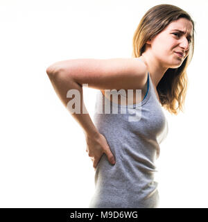 A young Caucasian woman girl wearing a grey vest, suffering from lower back pain, rubbing her back, standing against a white background, UK Stock Photo