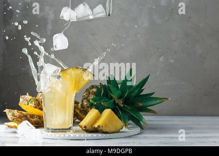 Pineapple fresh juice with falling ice cubes making splash. Cut fruit slices on wooden table and grey concrete background, front view Stock Photo
