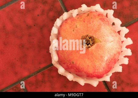 Whole red pomegranate in white frilly protective sleeve on red terracotta background, food still-life photograph Stock Photo