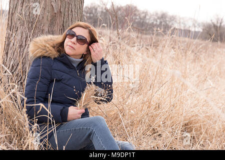 woman sitting alone by tree outside in field holding fresh picked bouquet of wildflowers Stock Photo