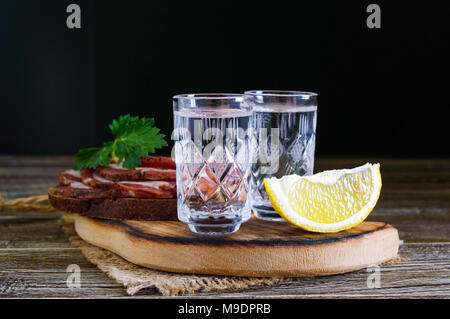 Two shot glasses of vodka with lemon slice and rye bread with salted bacon on the dark background. Traditional strong drink. Close-up. Stock Photo